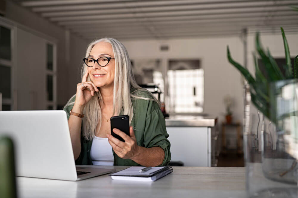 Happy senior woman holding smartphone and laptop daydreaming while looking away